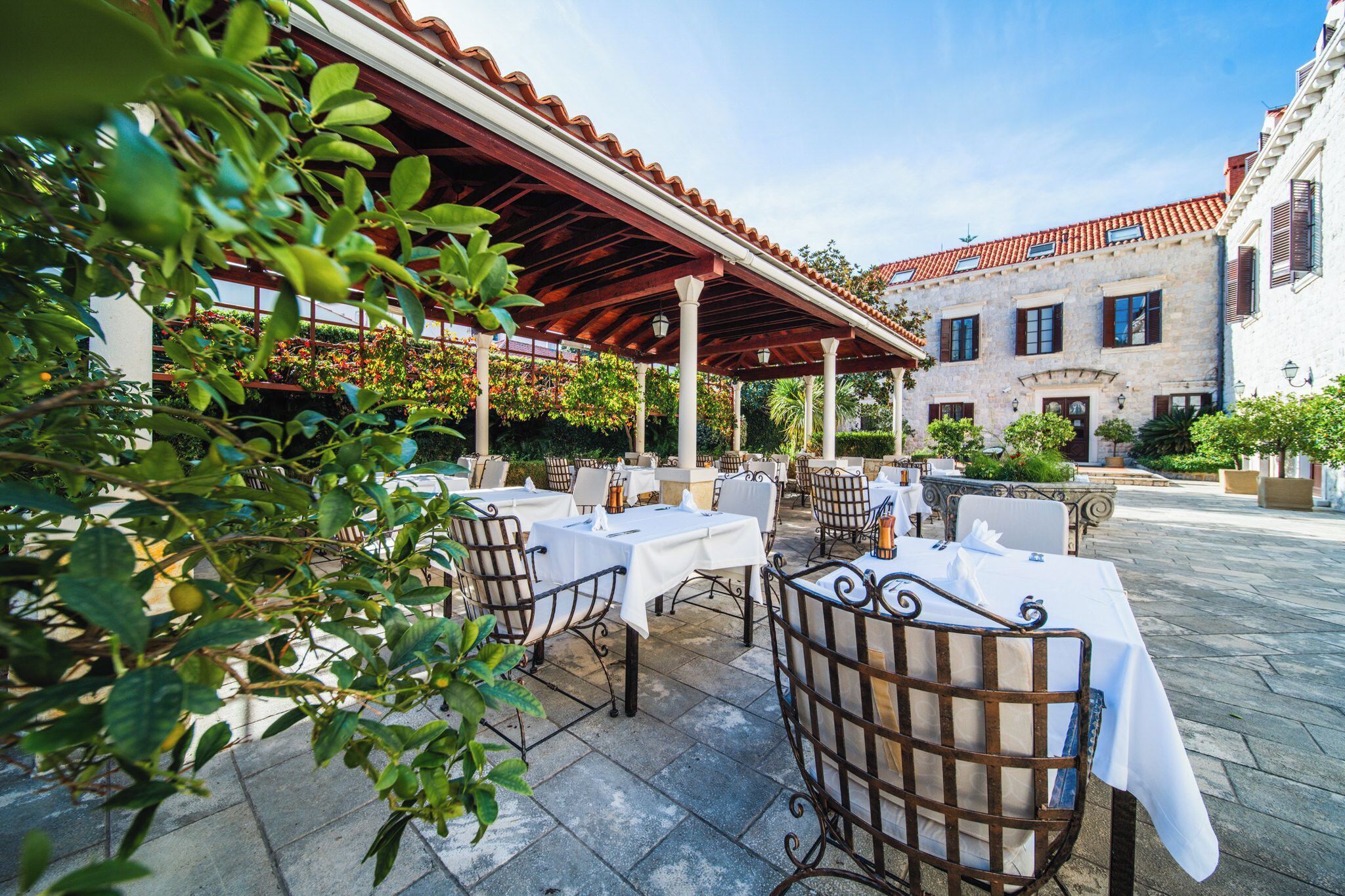 An outdoor patio with tables and chairs, a covered area, and a building with a tiled roof in the background.