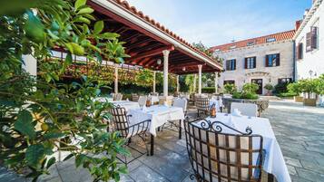 An outdoor patio with tables and chairs, a covered area, and a building with a tiled roof in the background.