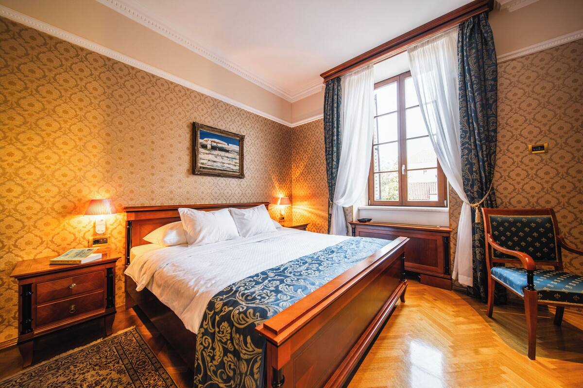 Ein Hotelzimmer mit einem großen Bett, zwei Nachttischlampen, einem Holznachttisch, einem Sessel, einem Fenster mit Vorhängen und einem Wandbild.