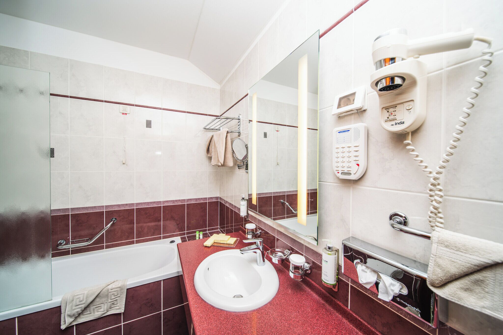 A modern bathroom with a white sink, red-tiled walls, a glass shower enclosure, a hairdryer, and a towel rack.