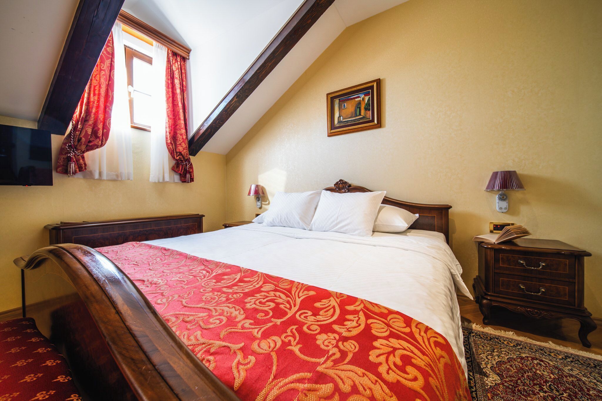 Quarto com uma cama grande, cabeceira de madeira, mesa de cabeceira com abajur, pequena mesa com um livro e um quadro na parede.