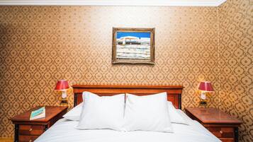 Uma cama bem arrumada com lençóis brancos, duas mesas de cabeceira com luminárias, um quadro na parede e papel de parede estampado.