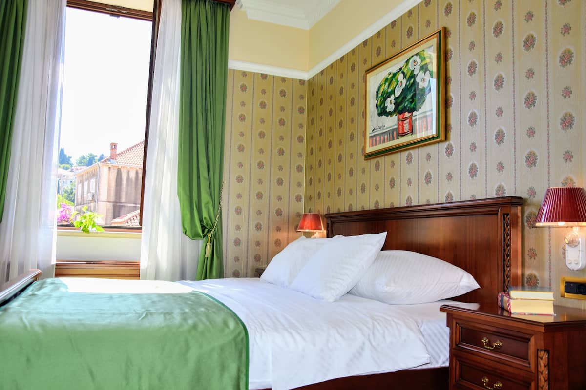 Une chambre d’hôtel avec un lit, une table de chevet, une lampe, un tableau encadré au mur et une fenêtre avec des rideaux.