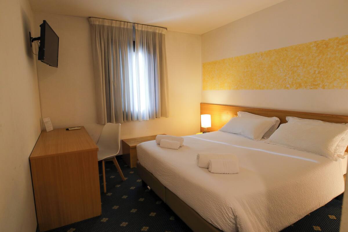 Een hotelkamer met een bed, een bureau, een stoel, een aan de muur bevestigde televisie en een raam met gordijnen.