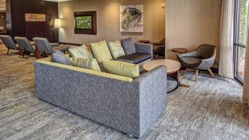 Un vestíbulo moderno con un sofá gris, sillones y una mesa redonda. Hay obras de arte en las paredes y una pantalla de televisión que muestra un partido deportivo.