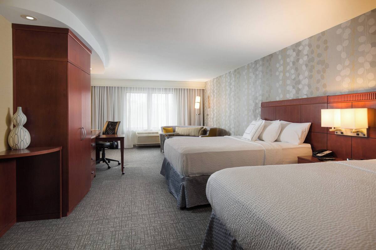 A hotel room with two beds, a desk, a chair, a lamp, and a large window with curtains.