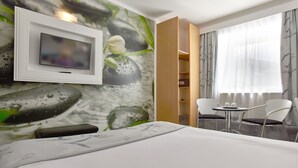 Habitación de hotel con cama, televisor montado en la pared, una mesita con sillas y ventana con cortinas.