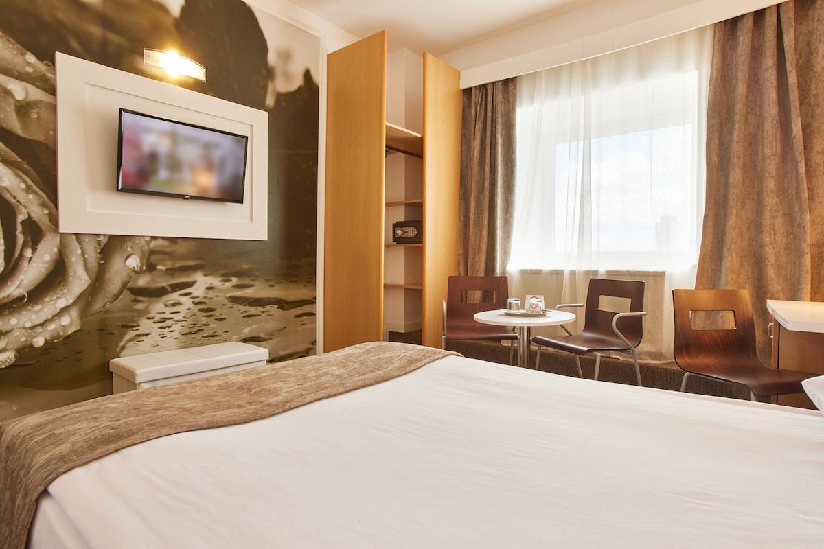 Une chambre d’hôtel avec un lit, un téléviseur fixé au mur, une petite table ronde avec des chaises et une fenêtre avec des rideaux.