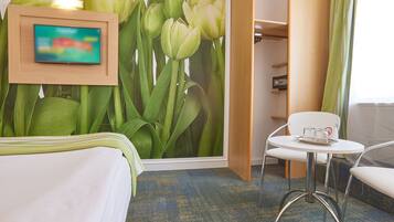A hotel room with a bed, a TV mounted on the wall, a table with a glass and a cup, and a chair.