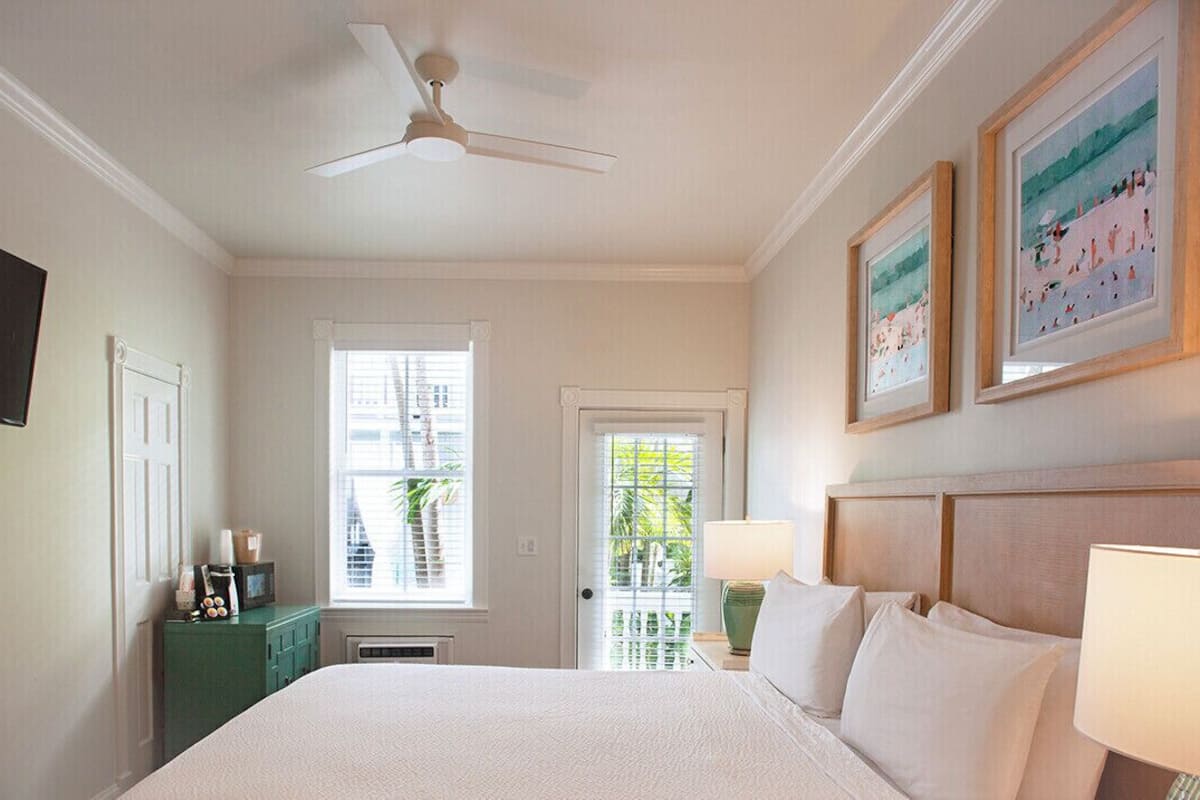 A bedroom with a bed, a ceiling fan, a window with blinds, a door, a green dresser, and framed artwork on the wall.