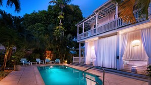 A swimming pool with lounge chairs, a covered deck, and a view of lush greenery. - Coco Plum Inn (Key West)
