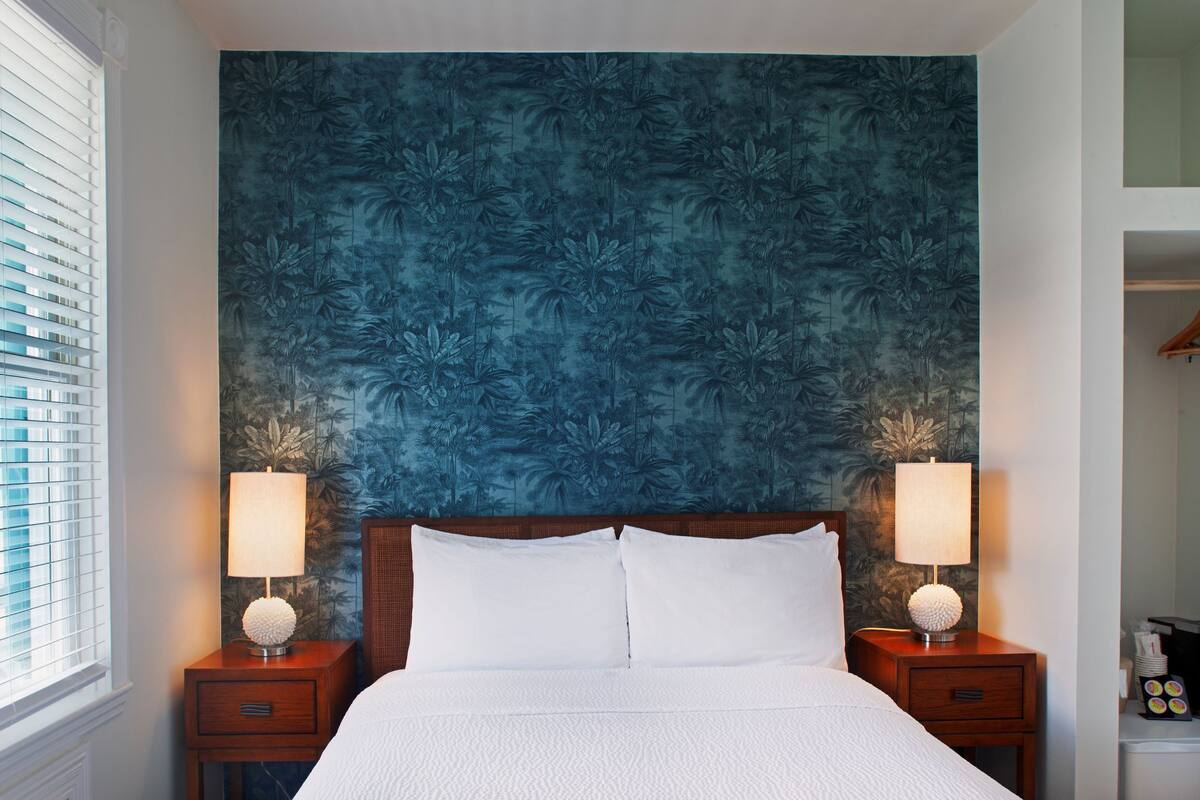 A hotel room with a large bed, two bedside tables with lamps, a nightstand with a lamp, and a window with blinds.