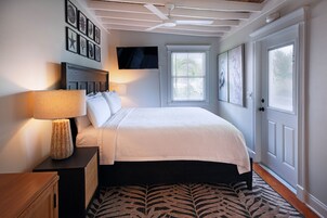Un dormitorio con cama, una mesita de noche con lámpara, ventilador de techo, ventana con persianas y cuadros en la pared.