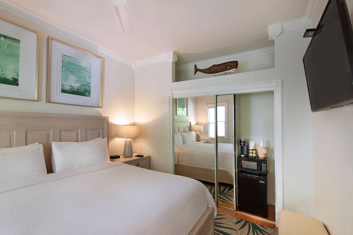 A hotel room with a large bed, a nightstand with a lamp, a wardrobe with a mirror, a flat-screen TV, and a small kitchenette.