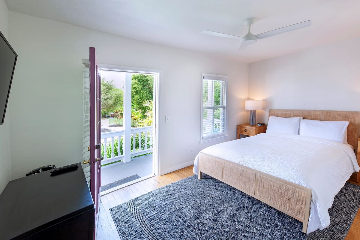A bedroom with a bed, a ceiling fan, a nightstand with a lamp, a door leading to a balcony, and a desk with a chair.