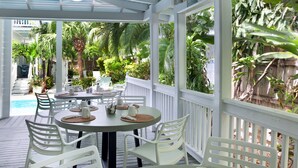 A covered outdoor dining area with white chairs and tables, surrounded by lush greenery and a wooden deck. - Coco Plum Inn (Key West)
