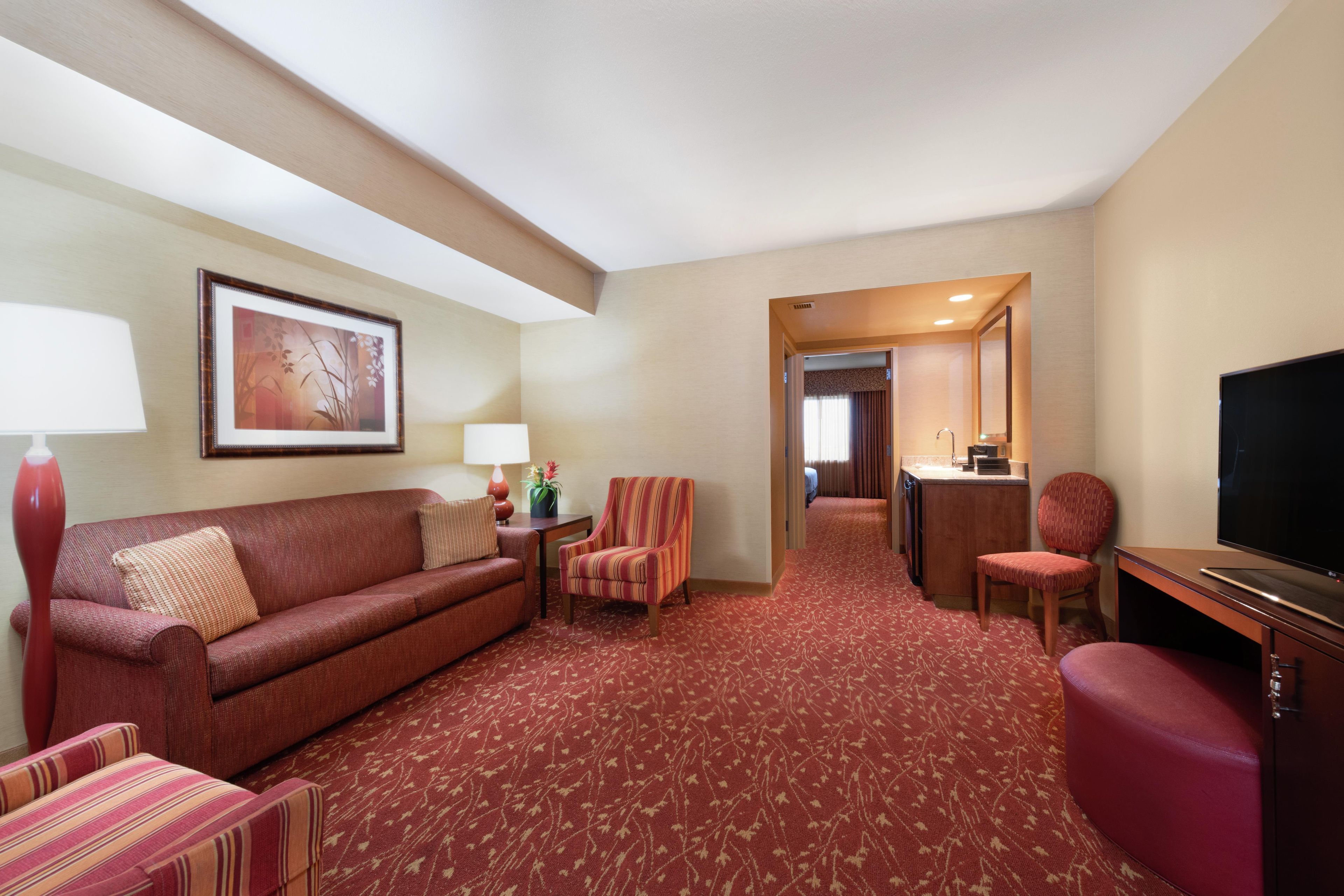 A hotel room with a red carpet, a sofa, a striped armchair, a desk with a television, and a bathroom visible through an open door.