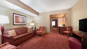 A hotel room with a red carpet, a sofa, a striped armchair, a desk with a television, and a bathroom visible through an open door.