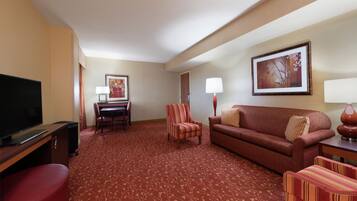 A hotel room with a television, a dining table with chairs, a sofa, a red patterned carpet, and a framed picture on the wall.