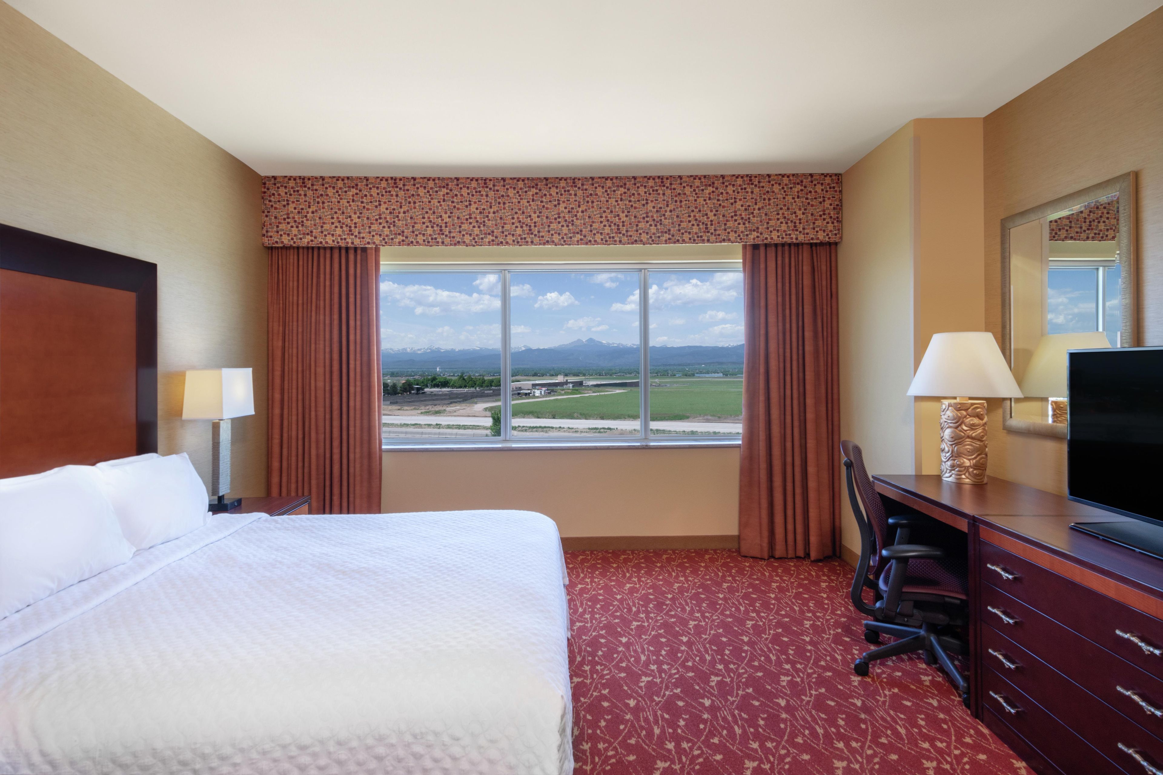 A hotel room with a bed, a desk, a television, and a large window with a view of the landscape.