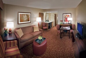 A hotel room with a living area featuring a sofa, ottoman, and a small table with a lamp. There is a television and a desk with a chair. - Embassy Suites by Hilton Loveland Conference Center (Loveland)