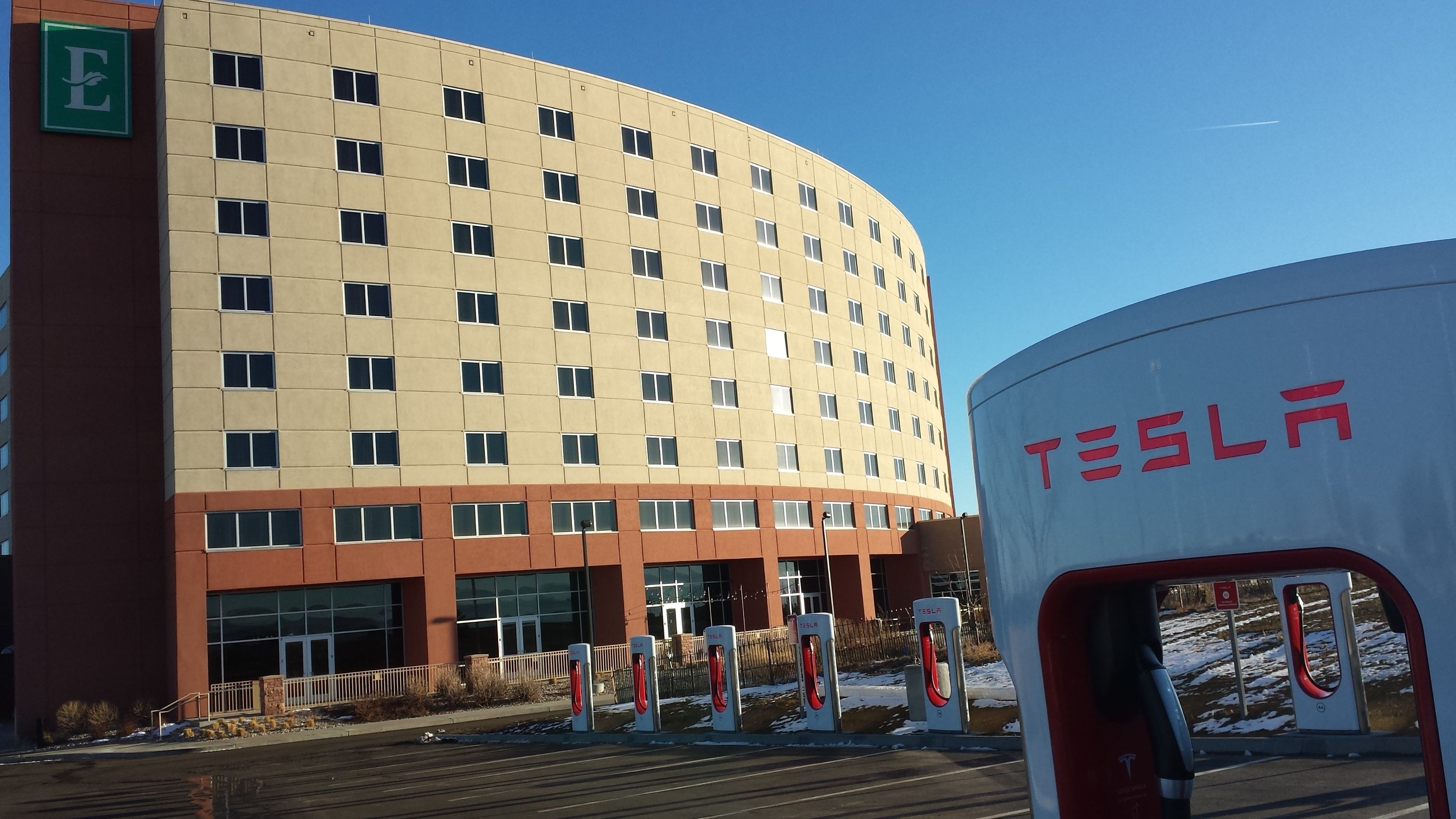 Un bâtiment moderne à la façade incurvée, doté de nombreuses fenêtres et d’une borne de recharge Tesla en premier plan.