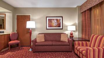 Habitación de hotel con sofá rojo, sillón a rayas y alfombra estampada. Se observa una puerta de madera, un cuadro en la pared y una lámpara sobre una mesita de noche.