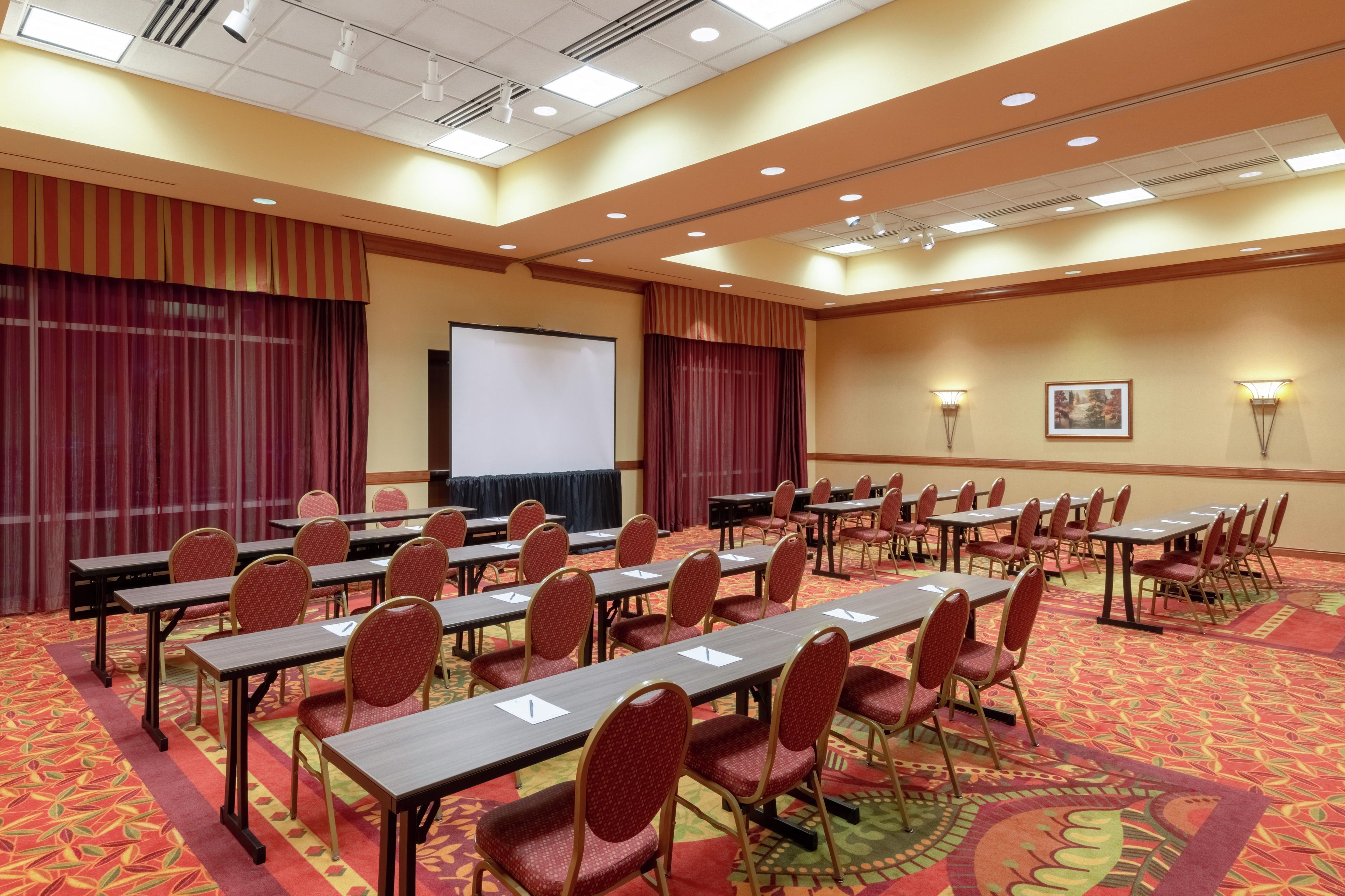 A conference room with rows of tables and chairs, a projection screen, and a carpeted floor.