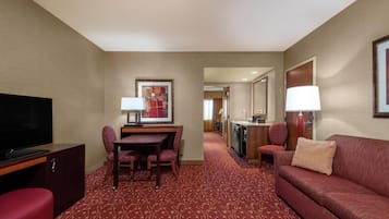 A hotel room with a flat-screen TV, a dining table with chairs, a sofa, a small kitchen area, and a hallway leading to another room.