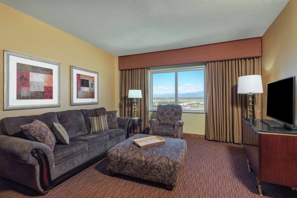 A hotel room with a large window, a sofa, a chair, a television, and framed artwork on the wall.