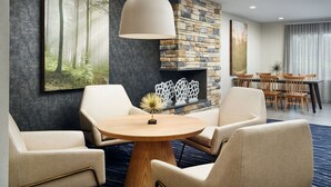 A modern hotel room with a round wooden table, beige armchairs, a stone fireplace, and a large forest-themed painting on the wall. - Fairfield Inn & Suites by Marriott Auburn Opelika (Opelika)