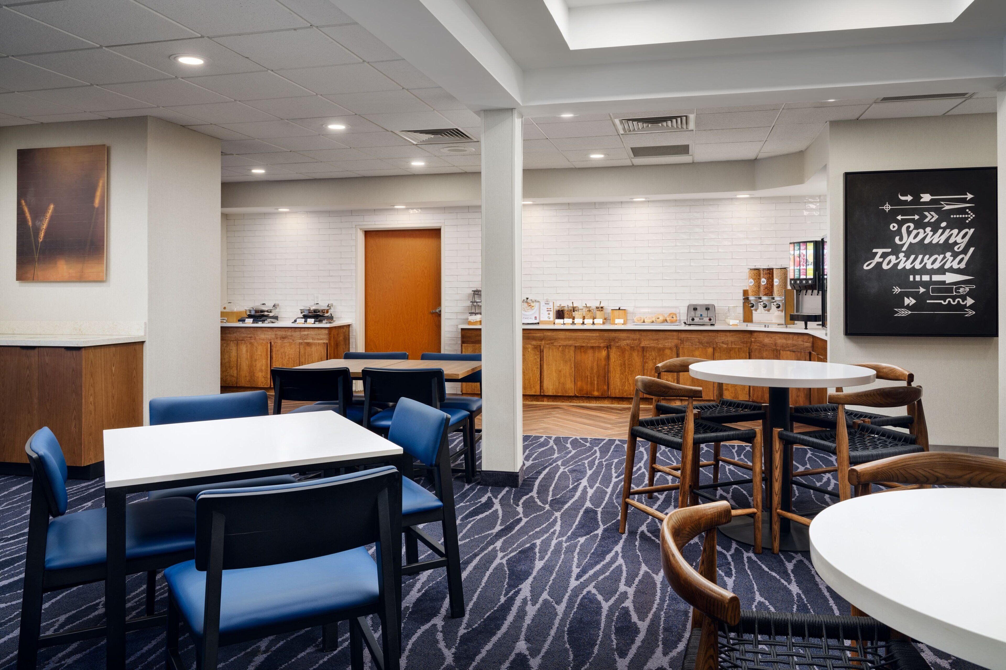 A hotel room with a dining area, a kitchen, and a bar. There are tables and chairs, a coffee machine, and a sign that says "Spring Forward".