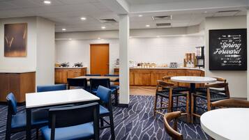 A hotel room with a dining area, a kitchen, and a bar. There are tables and chairs, a coffee machine, and a sign that says "Spring Forward".