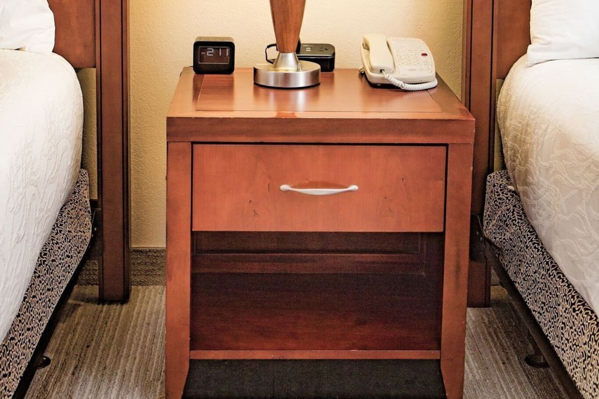 A hotel room with two beds, a bedside table with a lamp, a clock, and a phone.