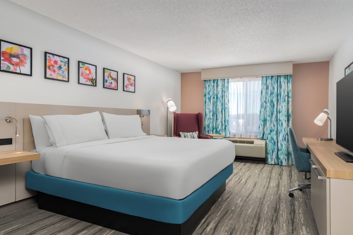 A hotel room with a large bed, a desk with a computer, a chair, a TV, and a window with curtains.
