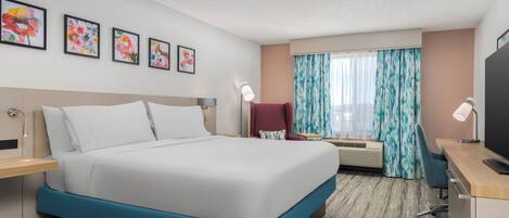 A hotel room with a large bed, a desk with a computer, a chair, a TV, and a window with curtains.