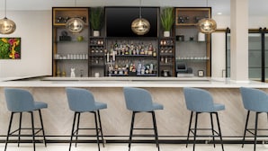 A modern bar area with three blue bar stools, a large selection of bottles and glasses on shelves, and pendant lighting.