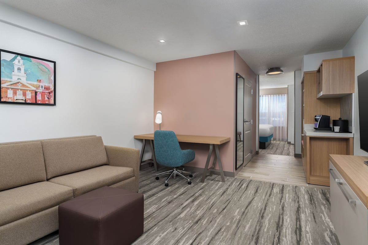 A modern hotel room with a sofa, a desk, a chair, a small ottoman, a kitchenette, and a painting on the wall.