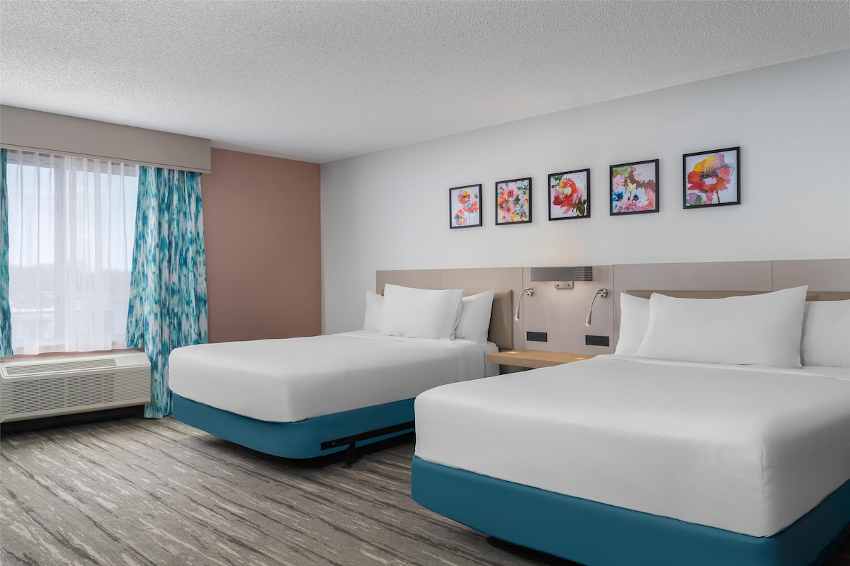 A hotel room with two beds, a window with curtains, a wall-mounted air conditioner, and four framed floral artworks.