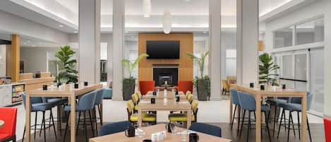 A spacious hotel lobby with multiple dining tables, comfortable seating, a large TV screen, and modern lighting fixtures.