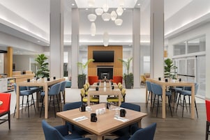 Un amplio vestíbulo de hotel con varias mesas de comedor, asientos cómodos, una pantalla de televisión grande y luminarias modernas.
