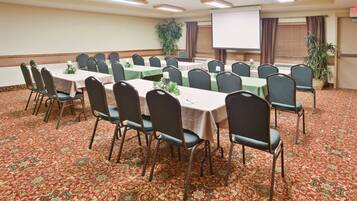 A conference room with multiple tables and chairs, a projector screen, and a large plant.