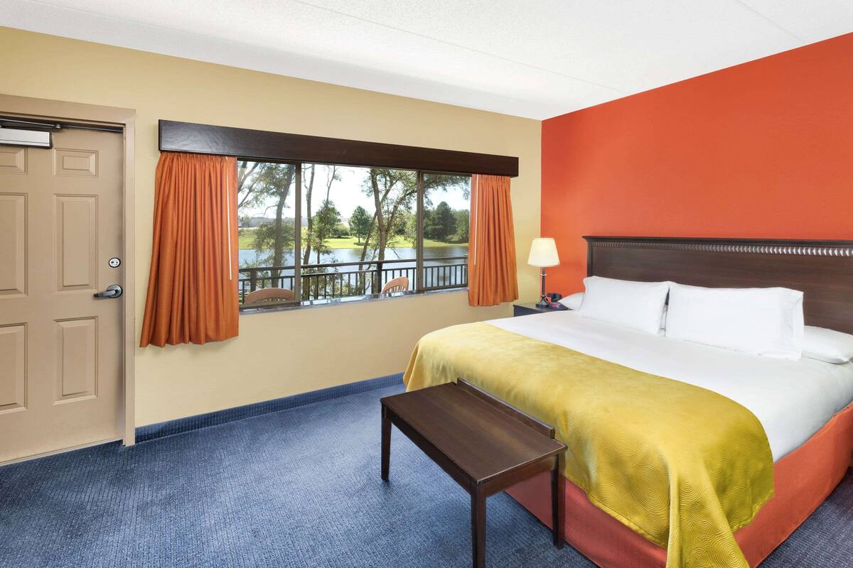 Una habitación de hotel con una cama, una mesita de noche, una lámpara, una ventana con cortinas y vistas a un lago.