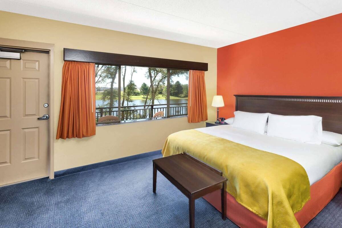 Una habitación de hotel con una cama, una mesita de noche, una ventana con cortinas, una puerta y vistas a un lago.