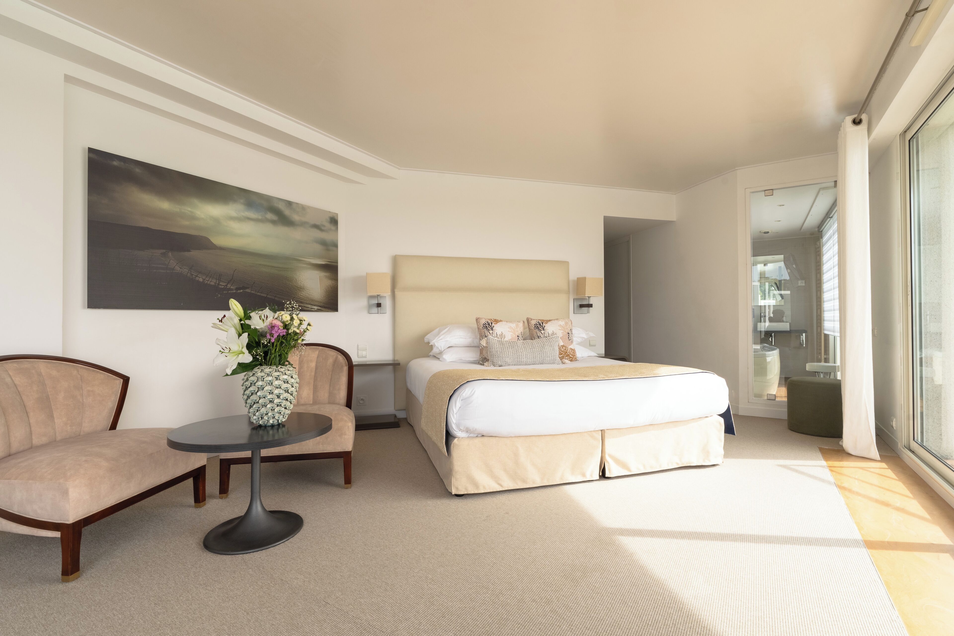 Habitación de hotel con una cama, una silla, una mesita con un florero y un cuadro en la pared.