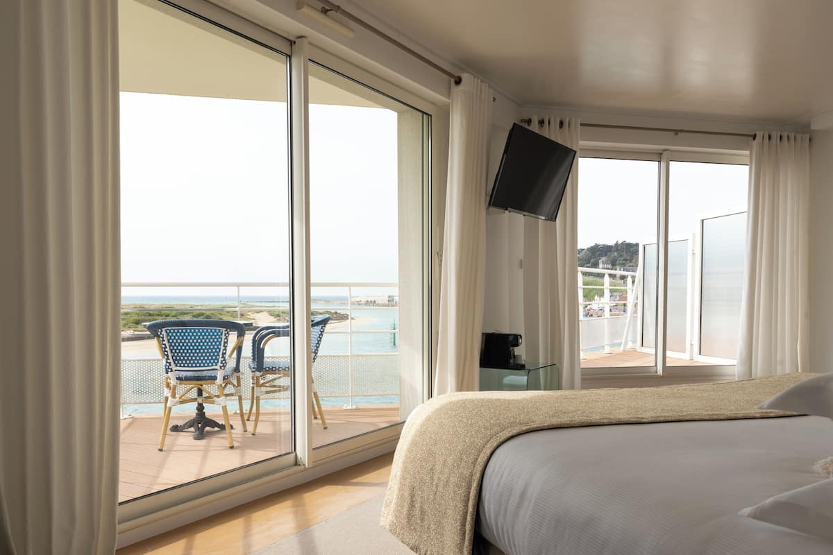 A hotel room with a large bed, a TV mounted on the wall, and a balcony with chairs and a view of the sea.