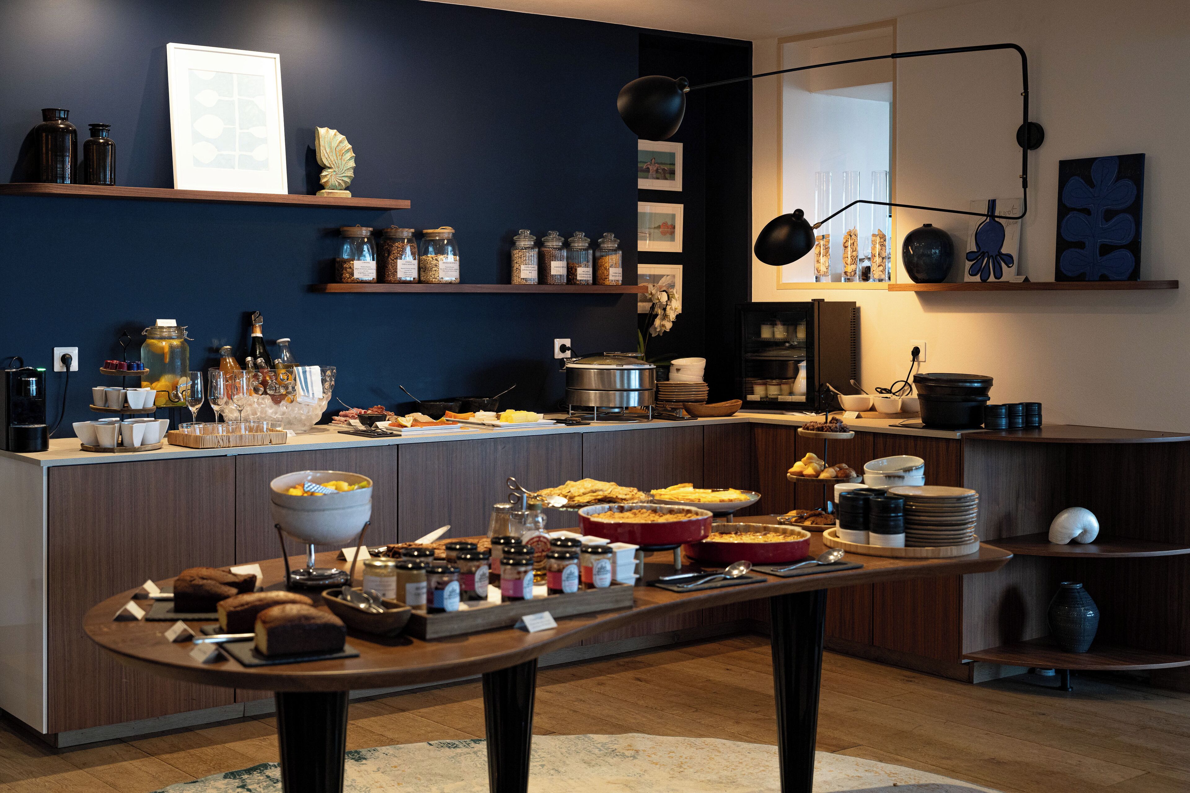 A buffet setup with various food items and condiments on a wooden table, surrounded by shelves with jars and kitchenware.
