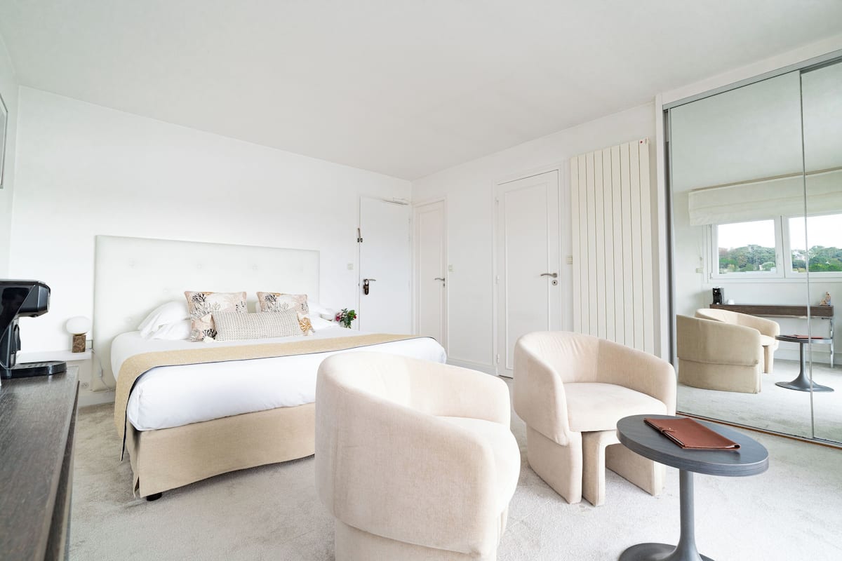A modern hotel room with a large bed, two armchairs, a small round table, and a large mirror reflecting the room.