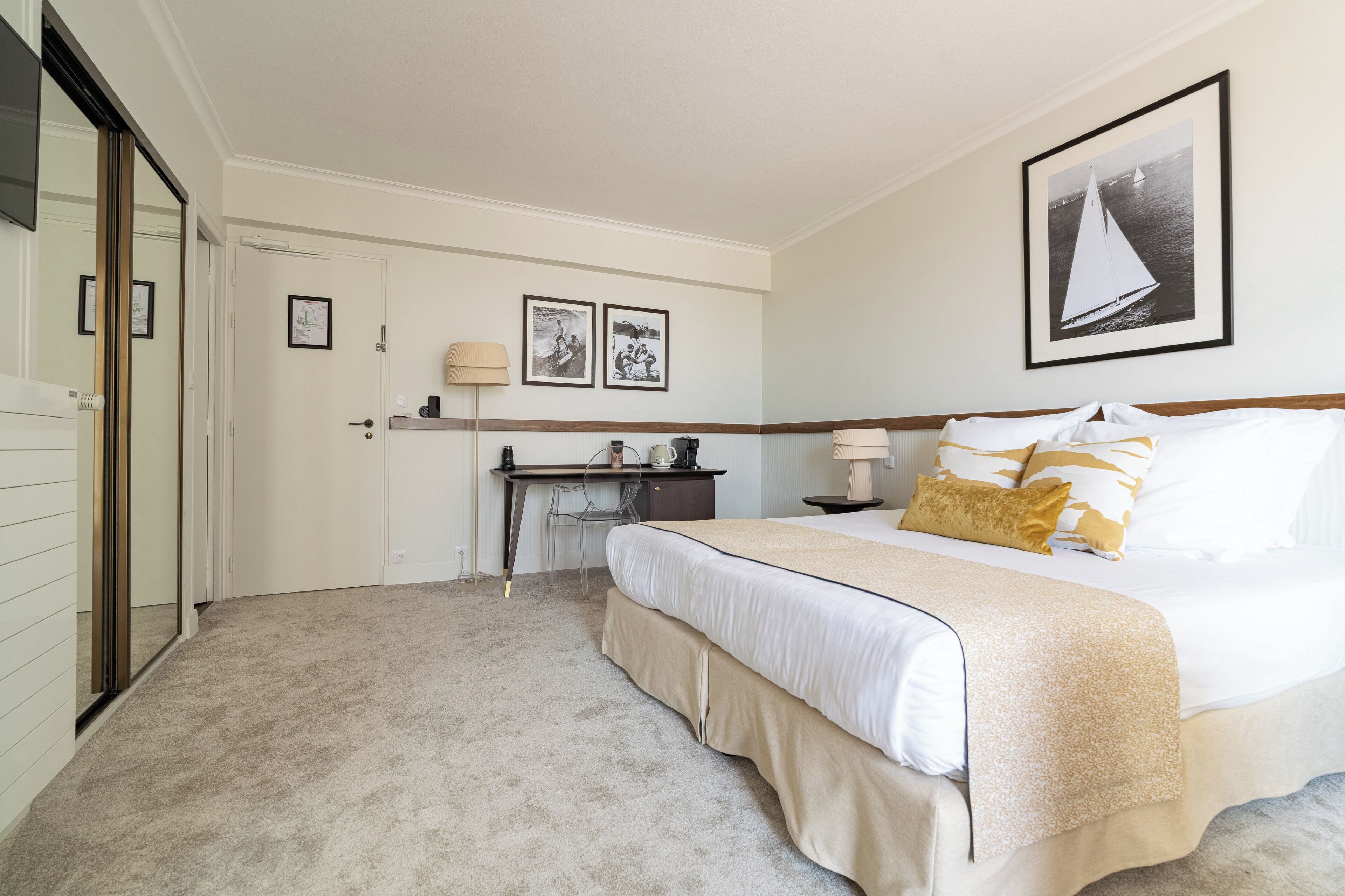 Ein Hotelzimmer mit Bett, Schreibtisch und Stuhl. An der Wand hängen Bilder in Rahmen und auf dem Schreibtisch steht eine Lampe.