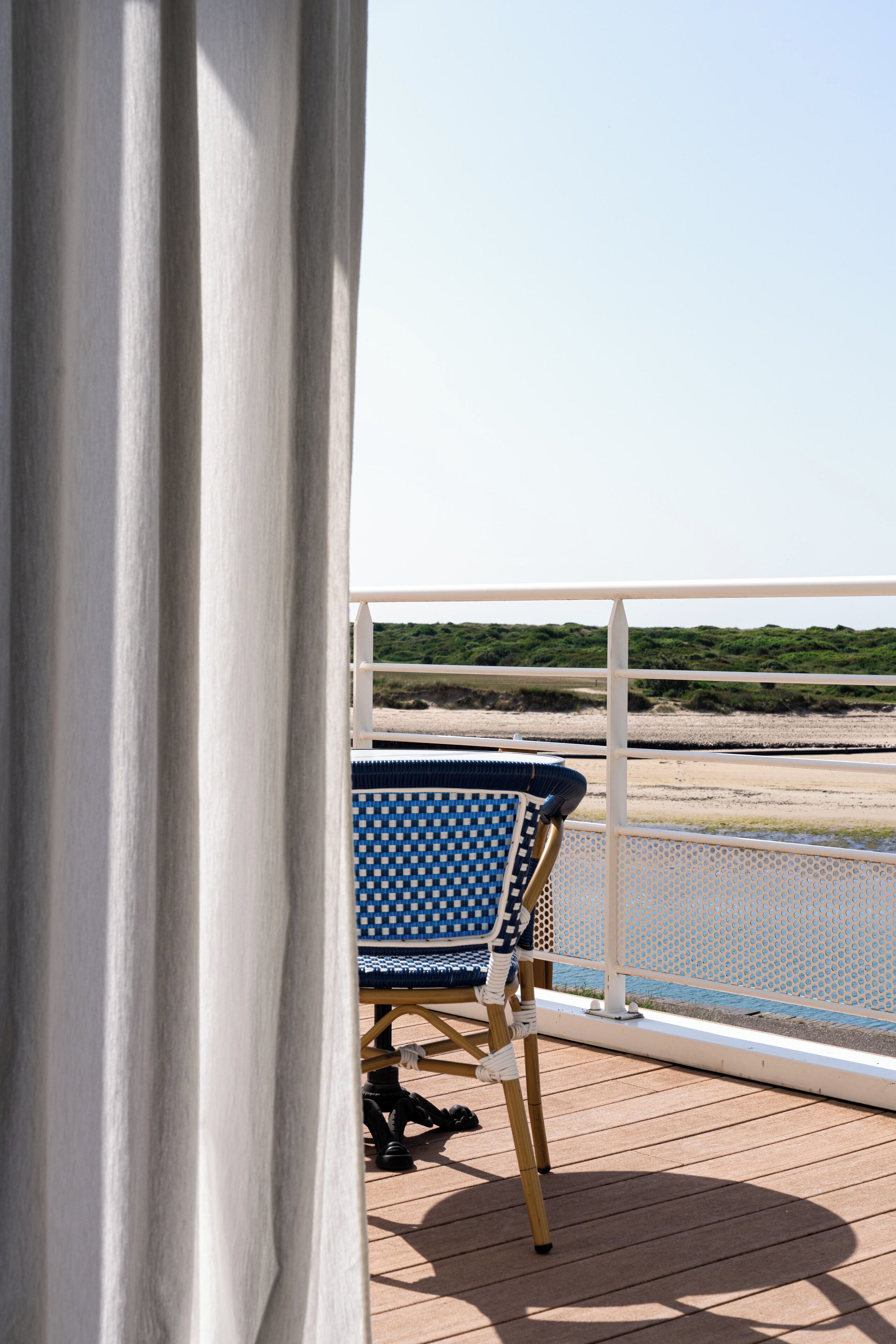 Un balcón con una silla azul y blanca a cuadros, barandilla blanca y vista a la playa y las dunas.